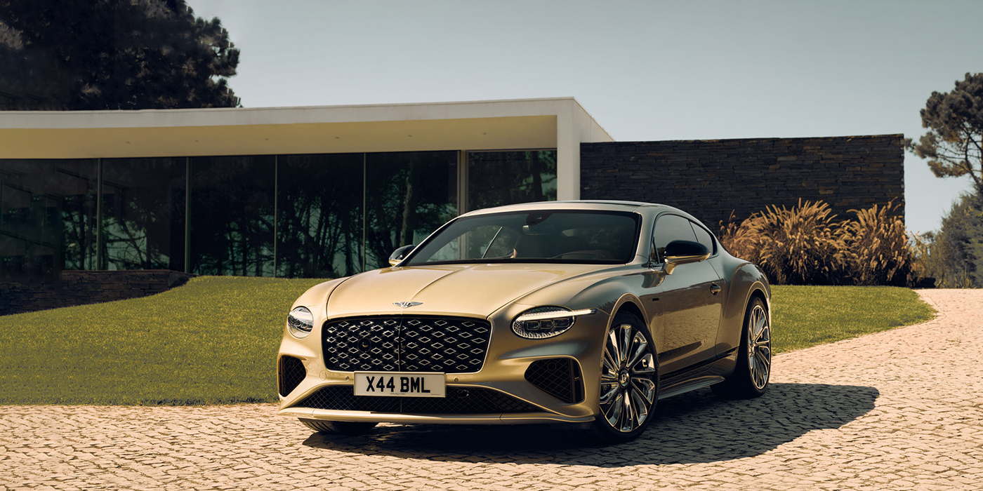 Bentley Belfast Bentley Continental GT Mulliner coupe front three quarters in Patina paint parked outside a glass fronted building with a grass lawn behind it
