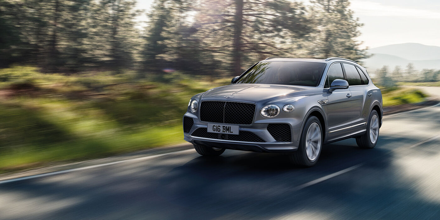 Bentley Belfast Bentley Bentayga luxury SUV front three quarter in Moonbeam paint, driving on an country road past trees.