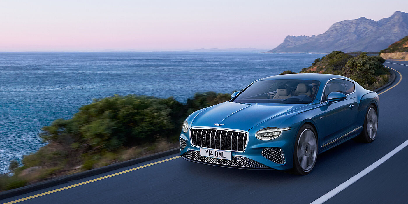 Bentley Belfast New Bentley Continental GT Azure coupe in Light Windsor blue paint with 22 inch Azure wheels and chrome grill, driving dynamically by the sea