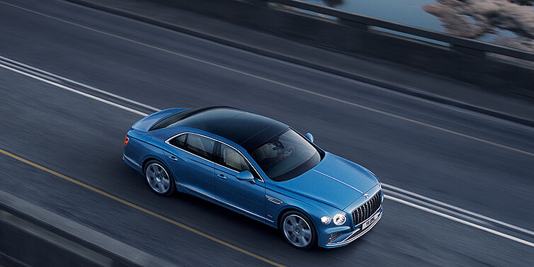 Bentley Belfast New Bentley Flying Spur Azure sedan in Light windsor blue paint by Mulliner, with 22 inch Azure wheels seen from overhead driving dynamically on a country road