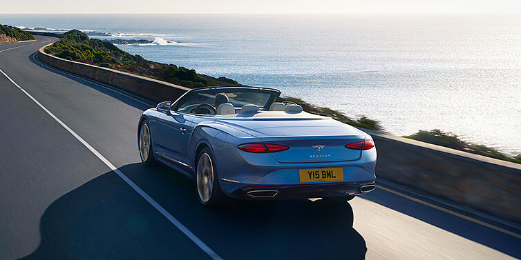 Bentley Belfast New Bentley Continental GTC Azure convertible rear view in Light Windsor blue paint with 22 inch ten spoke wheels driving dynamically on an ocean road in the sunshine
